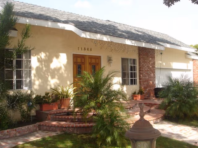 Front entrance of a beige senior living building with brick accents and potted plants.