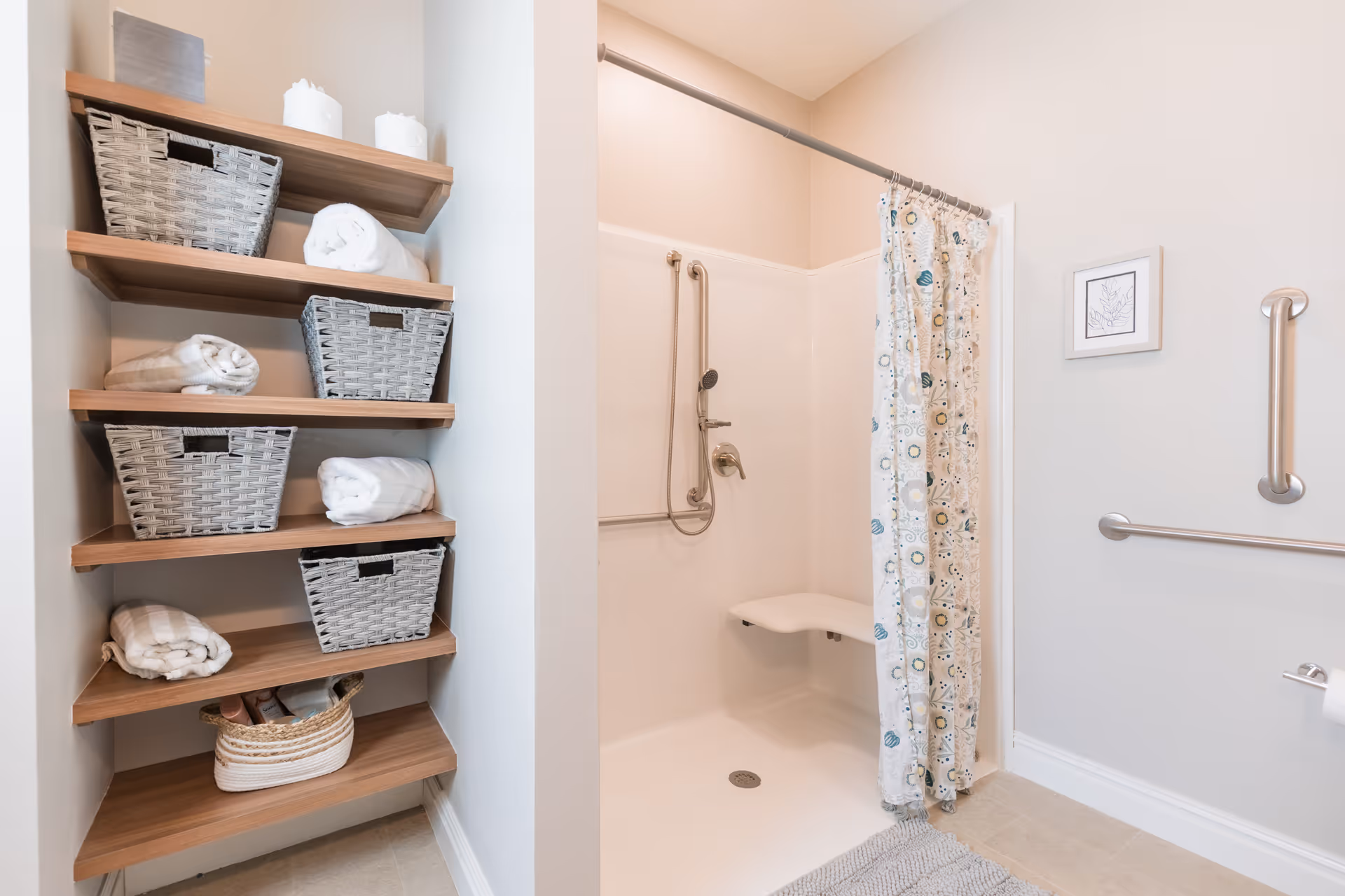A bathroom with a walk-in shower featuring a fold-down seat, grab bars, and a patterned shower curtain. To the left, there are wooden shelves holding woven baskets and neatly folded towels. The walls are light-colored, and there is a small framed picture on the wall next to the shower.