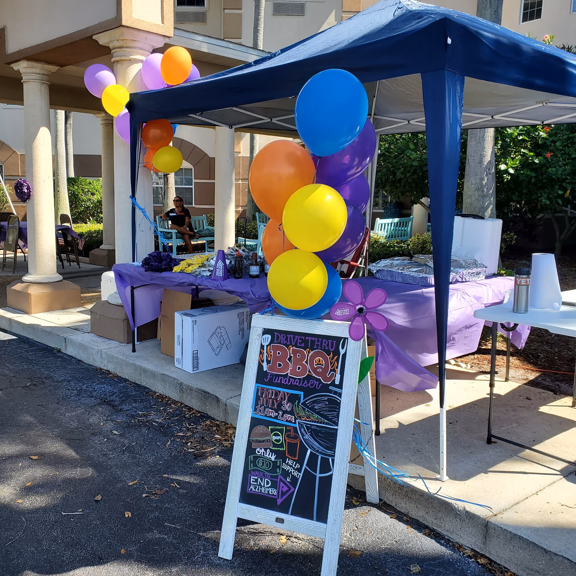 Outdoor fundraiser booth with balloons, a blue canopy, chalkboard sign near building entrance