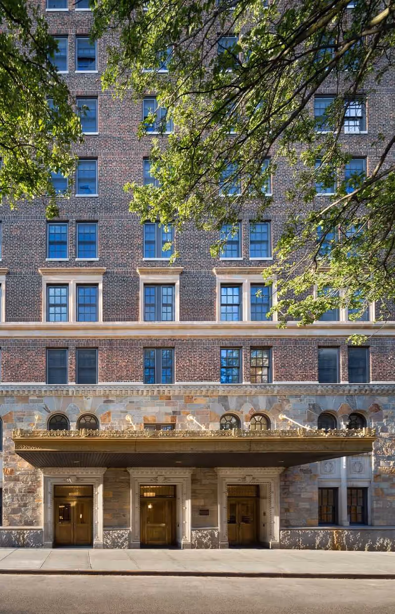 Brick building exterior with arched entrance, stone base, and leafy tree branches overhead.