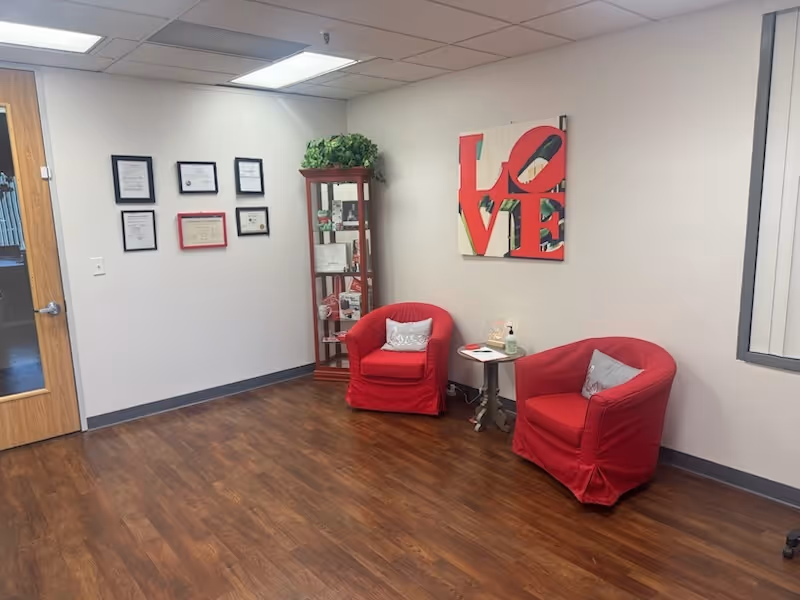 Interior lounge with two red chairs, a small table, plant, shelves, and wall art.