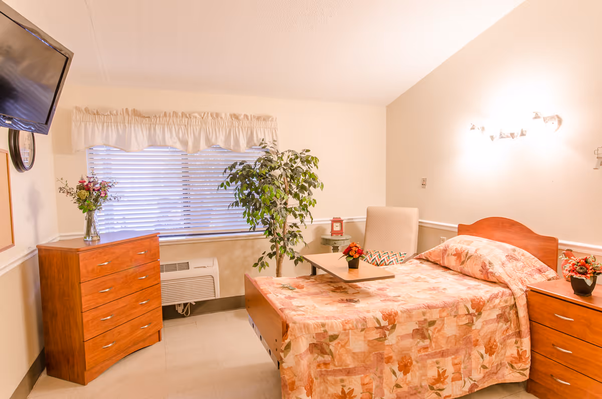 A cozy nursing center bedroom with a single bed covered in a floral bedspread, a wooden dresser with a vase of flowers, a window with blinds and a valance, a potted plant, a comfortable chair with a small table in front, and a wall-mounted television.