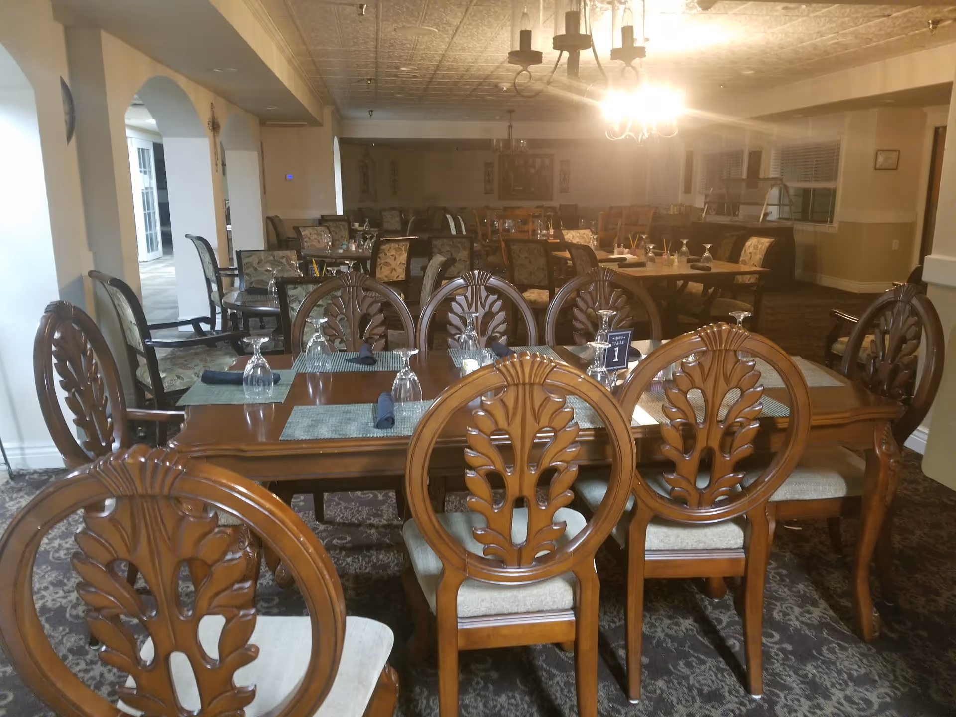 Empty dining room with wooden tables, ornate chairs, and place settings under warm chandelier lighting.
