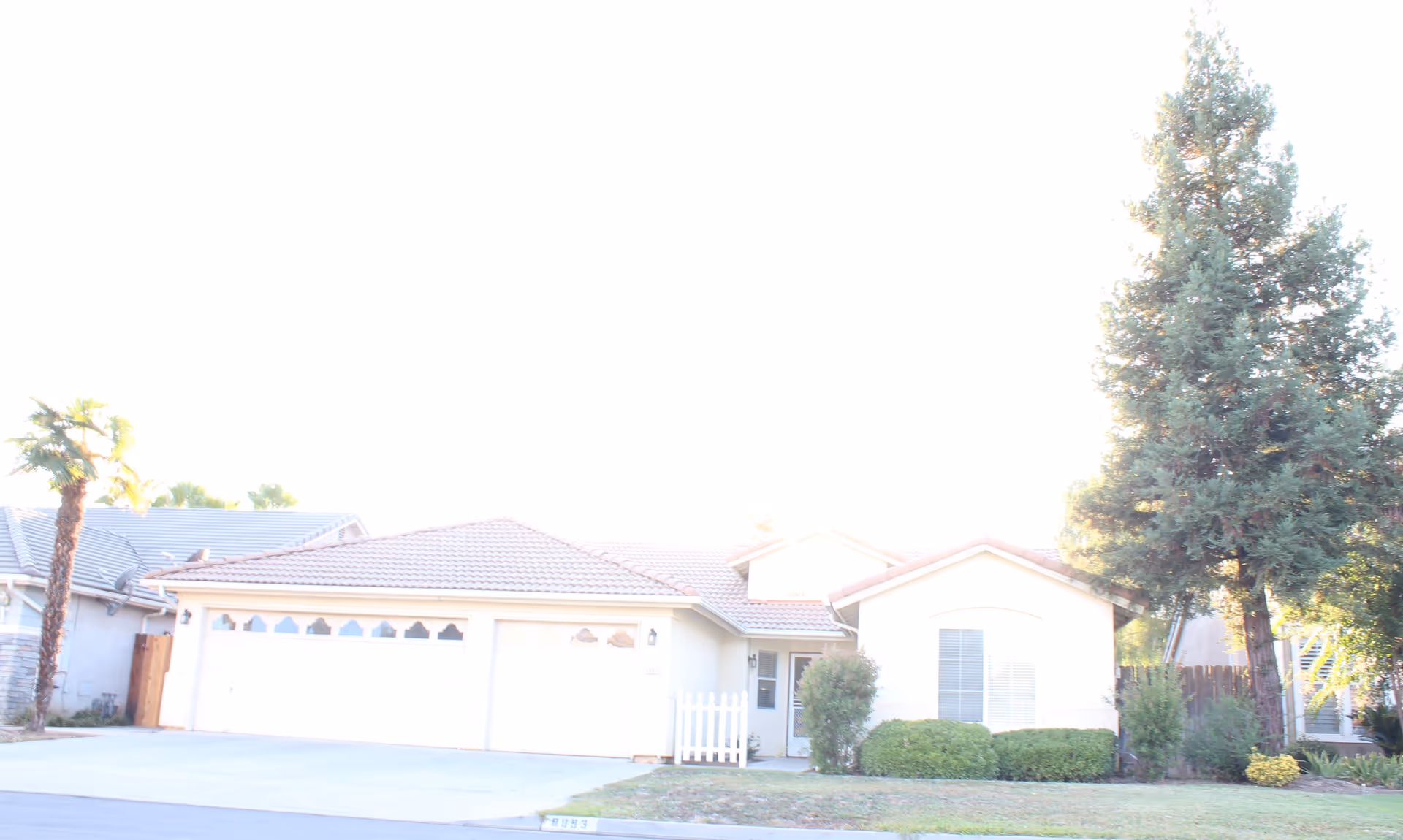 Front view of a single-story house with an attached three-car garage, driveway, lawn, and a tall tree on the right.