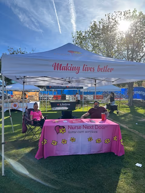 Outdoor event booth under a white tent with a pink tablecloth and two people seated.