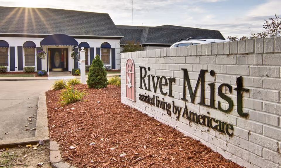 A brick entrance sign reading 'River Mist assisted living by Americare' sits in front of a landscaped driveway and the facility building with blue awnings.