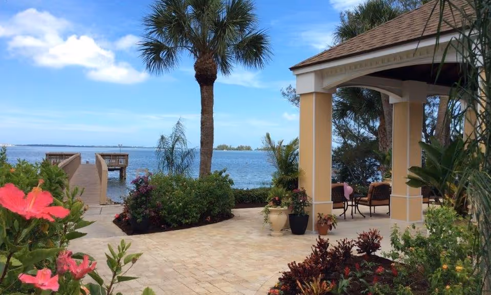 Landscaped waterfront patio with a covered gazebo, seating, potted plants, and a wooden dock extending into the water.