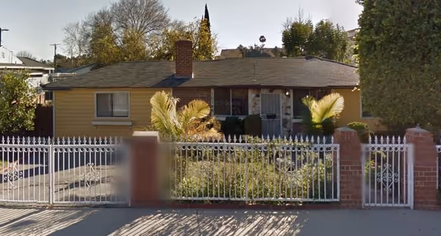 Single-story yellow house with a front yard, palm plants and a white metal fence facing the street.