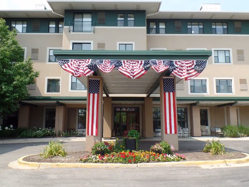 Front entrance of a senior residence building with a covered porte-cochere decorated with American flags and flower beds.