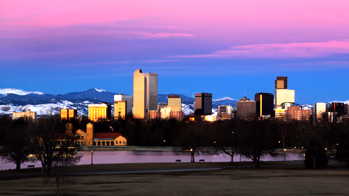 City skyline at sunset with mountains, river, and park trees in the foreground.