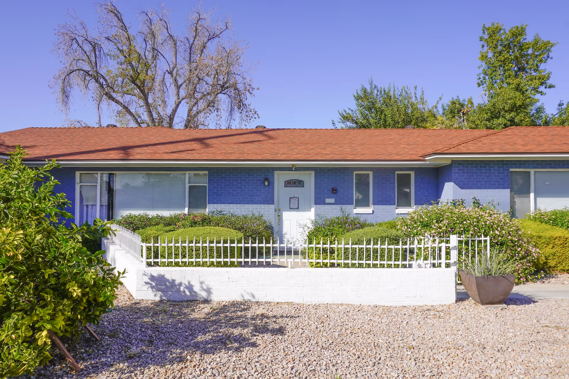 Front exterior view of a single-story assisted living home with blue painted brick walls, a red shingle roof, white front door, and white fence enclosing a small garden with bushes and plants. The sky is clear and blue.