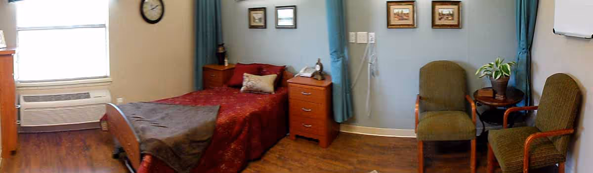 A cozy bedroom in a nursing facility featuring a single bed with red bedding and a gray blanket, two wooden nightstands with a clock and telephone, a window with blue curtains, a wall clock, two framed pictures on the wall, and two green upholstered chairs with wooden arms next to a small round table holding a potted plant.