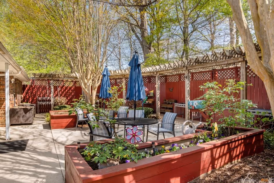 Outdoor patio area with raised red wooden garden beds filled with plants and flowers, round black metal table with four chairs, two closed blue umbrellas, and a wooden pergola along a red fence. Trees with green leaves provide shade and sunlight filters through.