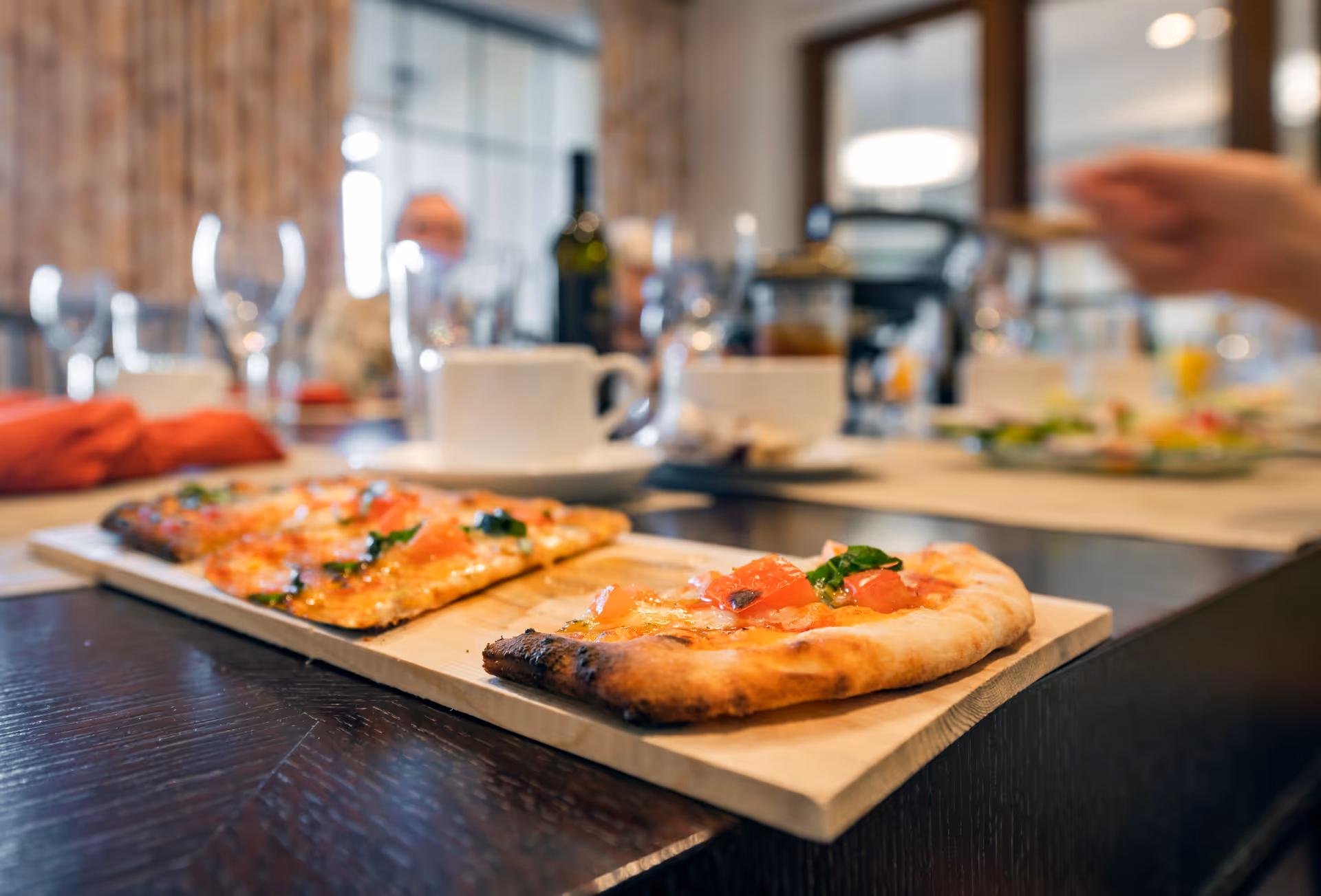 Close-up view of two slices of pizza on a wooden serving board on a dining table, with blurred background showing cups, glasses, a bottle, and people seated around the table in a dining room setting.