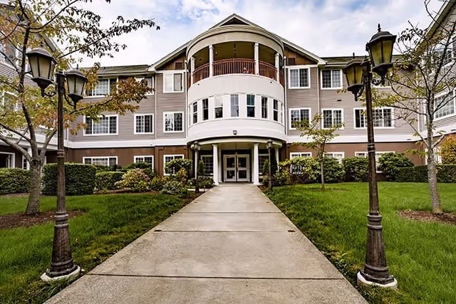 Front exterior view of a multi-story senior living facility building with a central rounded balcony, large windows, and a walkway leading to the entrance. The building is surrounded by green grass, trees, and vintage-style street lamps.