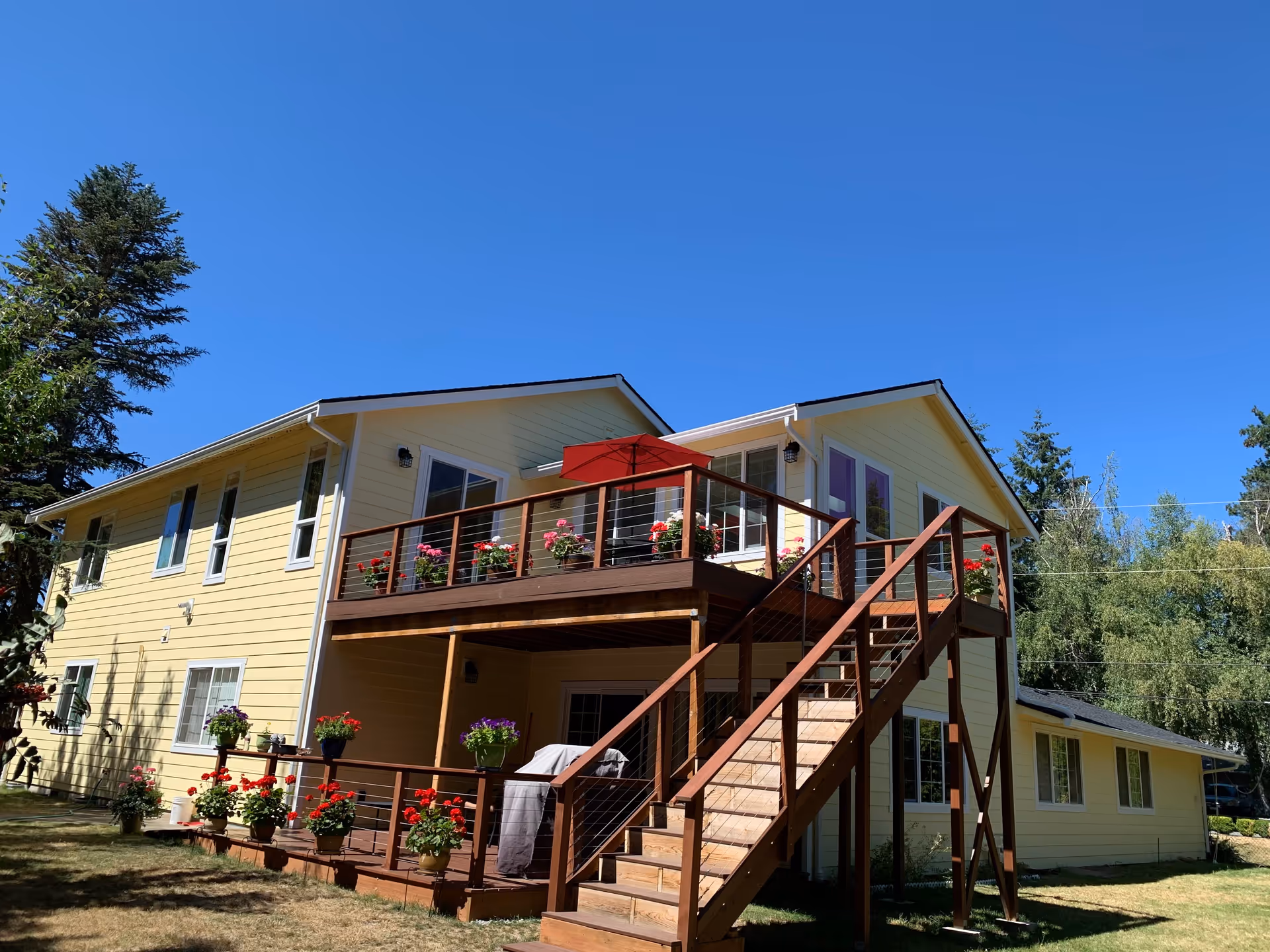 Exterior view of a two-story yellow building with a wooden deck and stairs leading to the upper level. The deck has flower pots with colorful flowers and a red umbrella. The building is surrounded by trees and a clear blue sky is visible.
