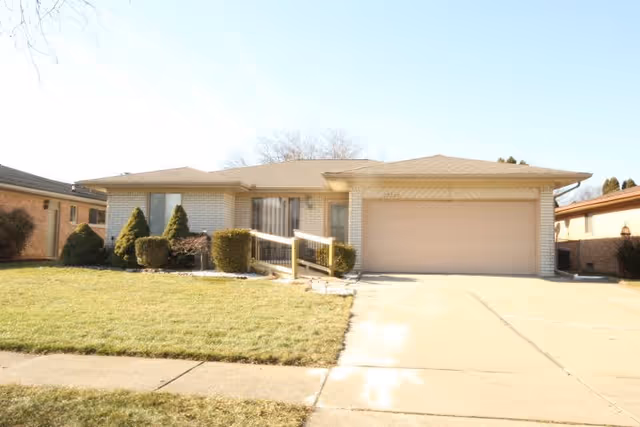 Single-story brick ranch house with a two-car garage, concrete driveway, front lawn, shrubs, and a small ramp leading to the entrance.