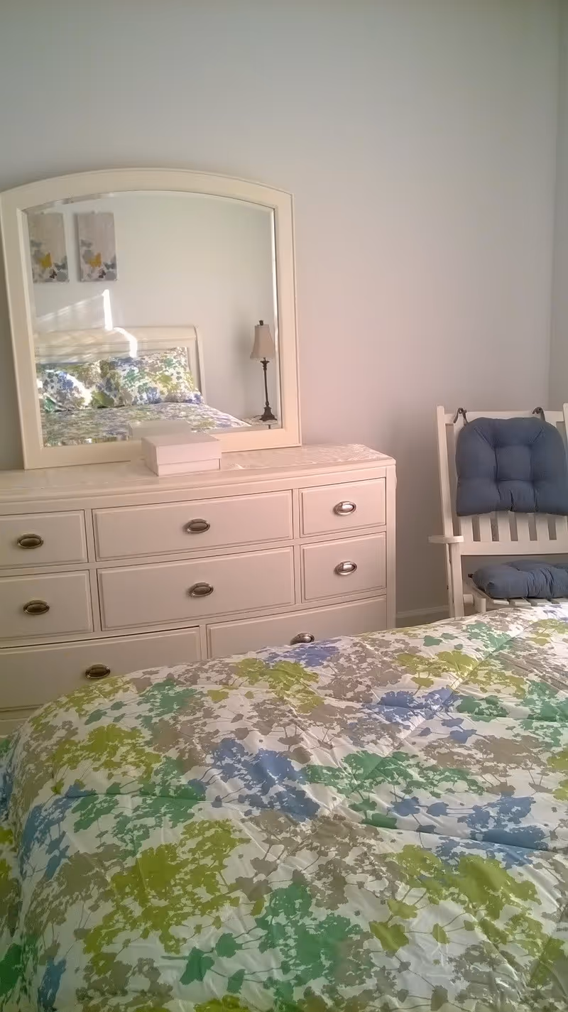 A bedroom with a white dresser featuring six drawers and a large mirror on top. The mirror reflects a bed with a floral patterned bedspread in shades of green, blue, and gray. A white rocking chair with blue cushions is positioned next to the dresser. The walls are light-colored and there is a lamp visible in the mirror reflection.