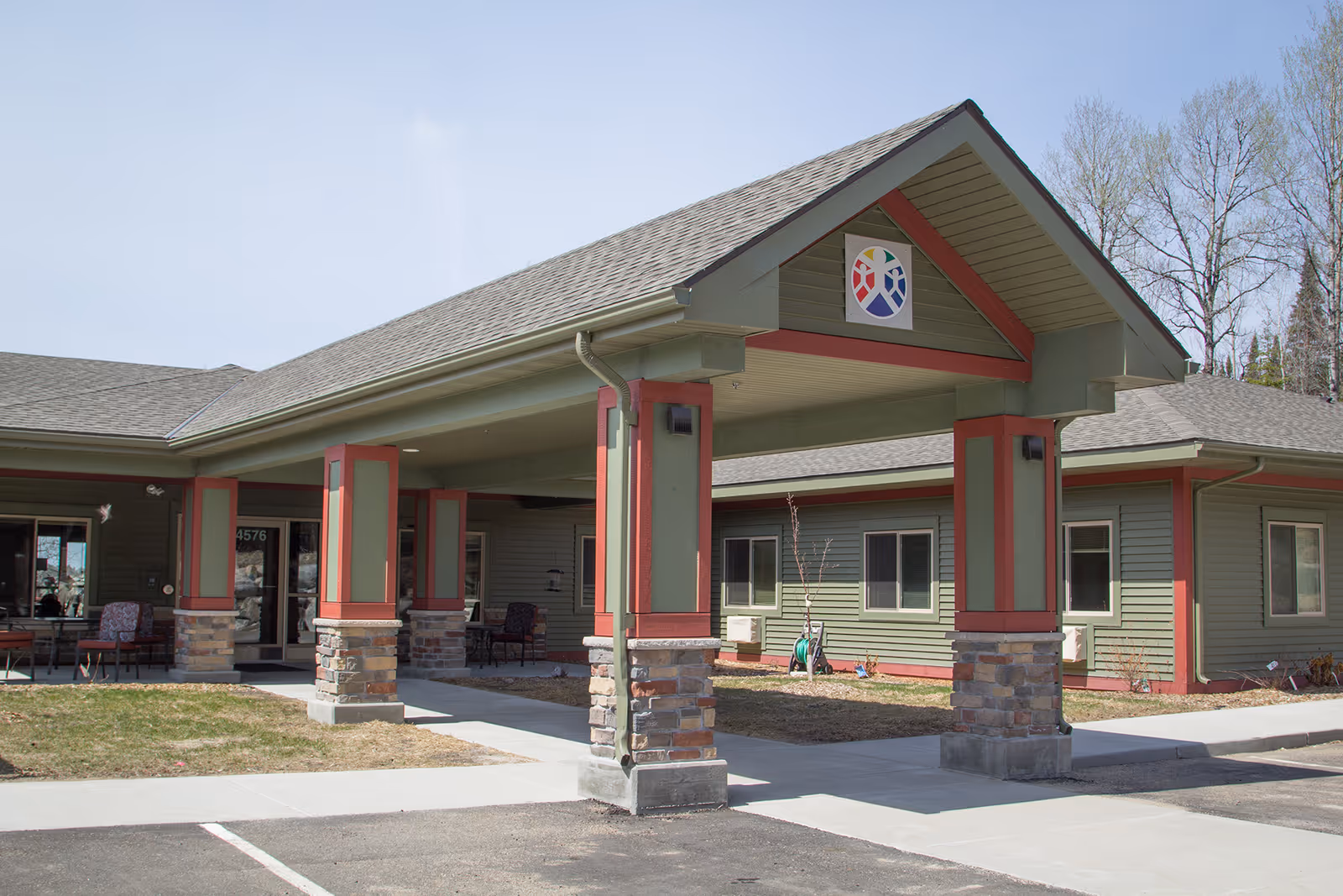 Exterior view of Carefree Living Orr facility entrance with a covered drop-off area supported by columns with stone bases and green and red accents. The building has green siding and multiple windows, with a logo displayed on the front gable. There are some chairs placed near the entrance and a paved driveway and sidewalk leading to the door.