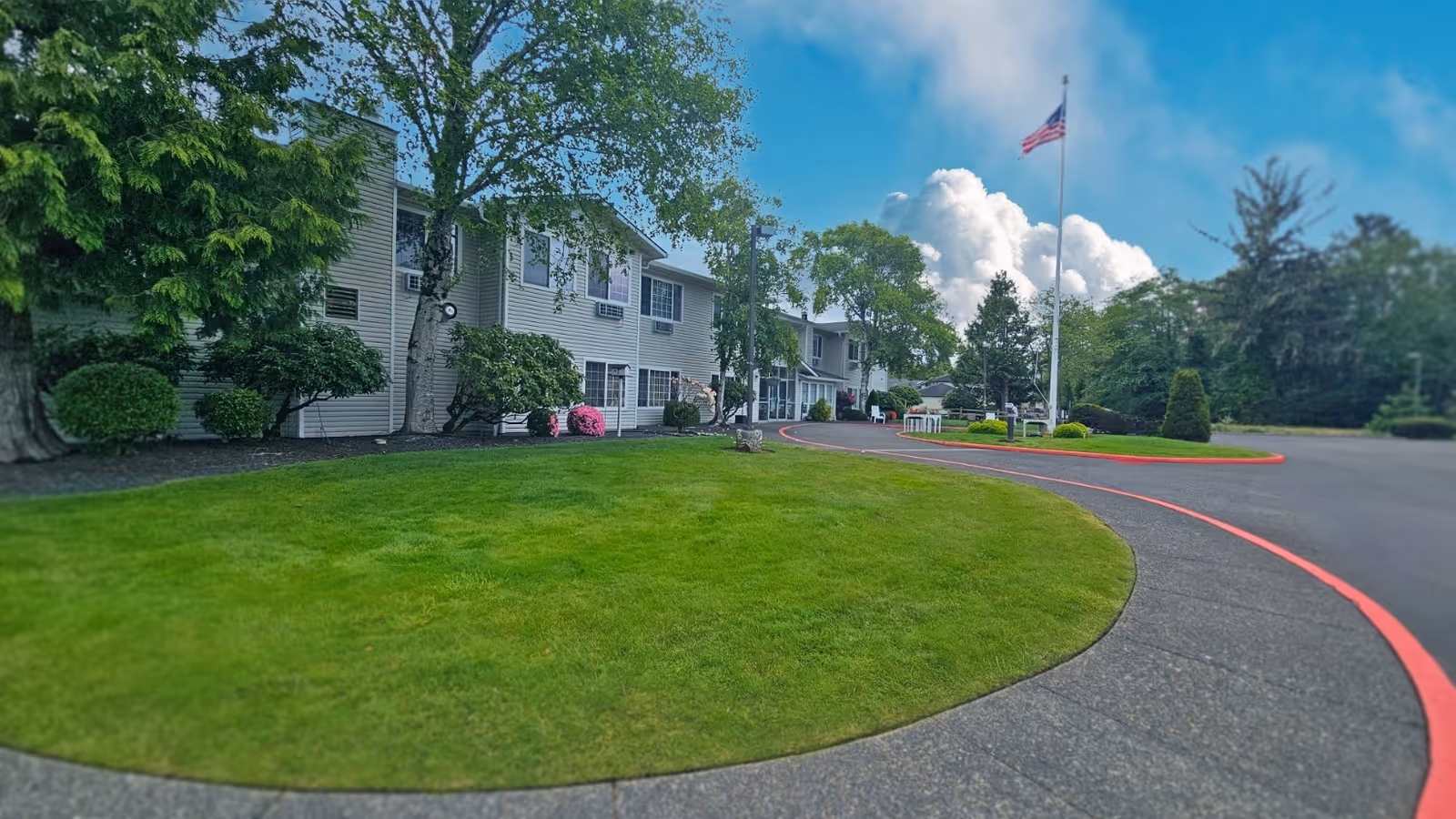 Front exterior of a two-story senior living facility with a curved driveway, manicured lawn, trees, and a flagpole.