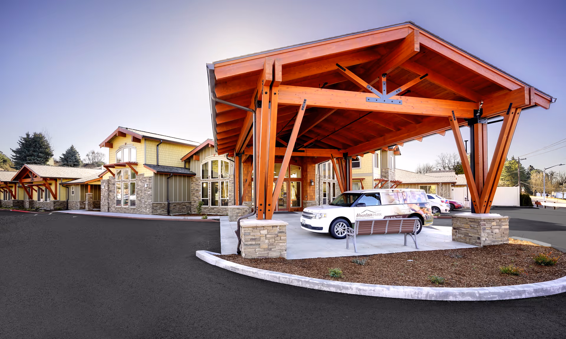 Exterior view of Mountain Park Memory Care facility featuring a large wooden covered entrance with stone pillars, a white vehicle parked under the entrance, and a bench nearby. The building has multiple windows and a combination of stone and siding on the walls. Trees and a clear sky are visible in the background.