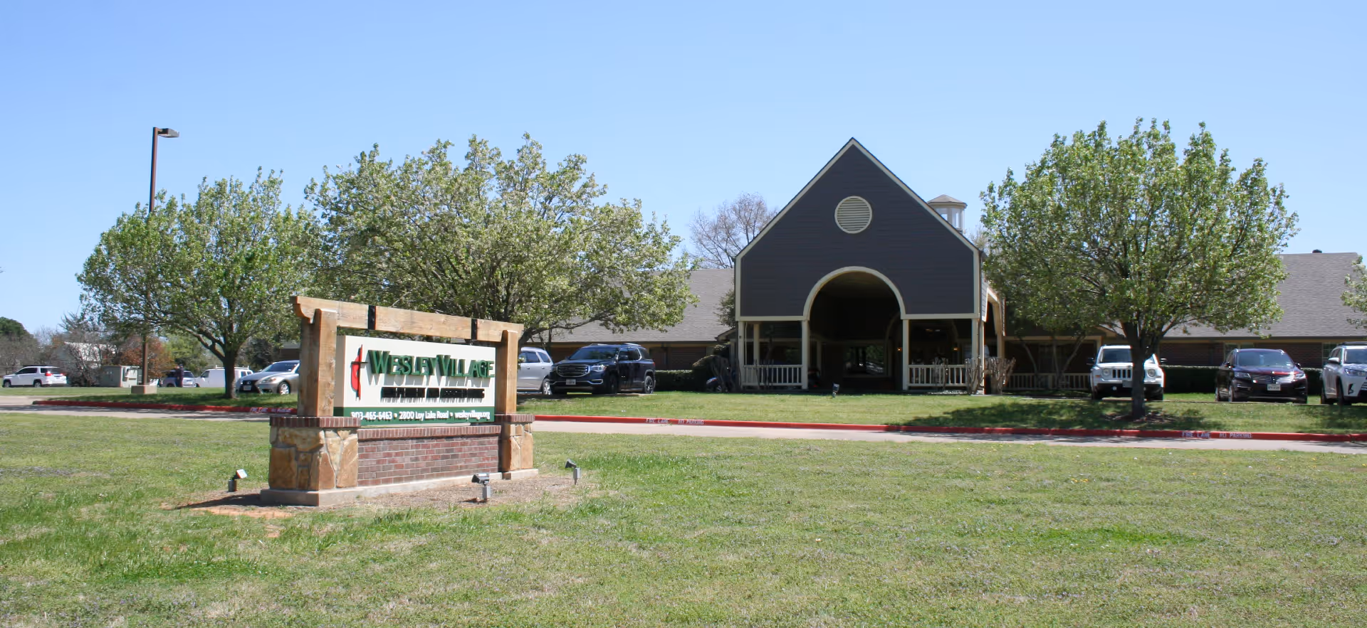 Exterior view of Wesley Village senior living facility with a large green lawn in front, a wooden and brick sign displaying the facility's name, several trees, parked cars, and a building with a covered entrance under a clear blue sky.
