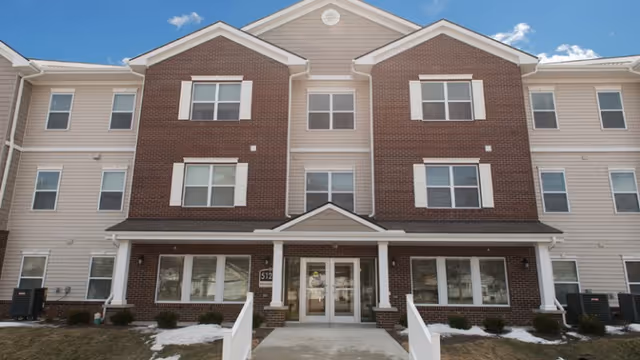 Three-story brick-and-beige senior living building with central glass doors and a covered entrance.
