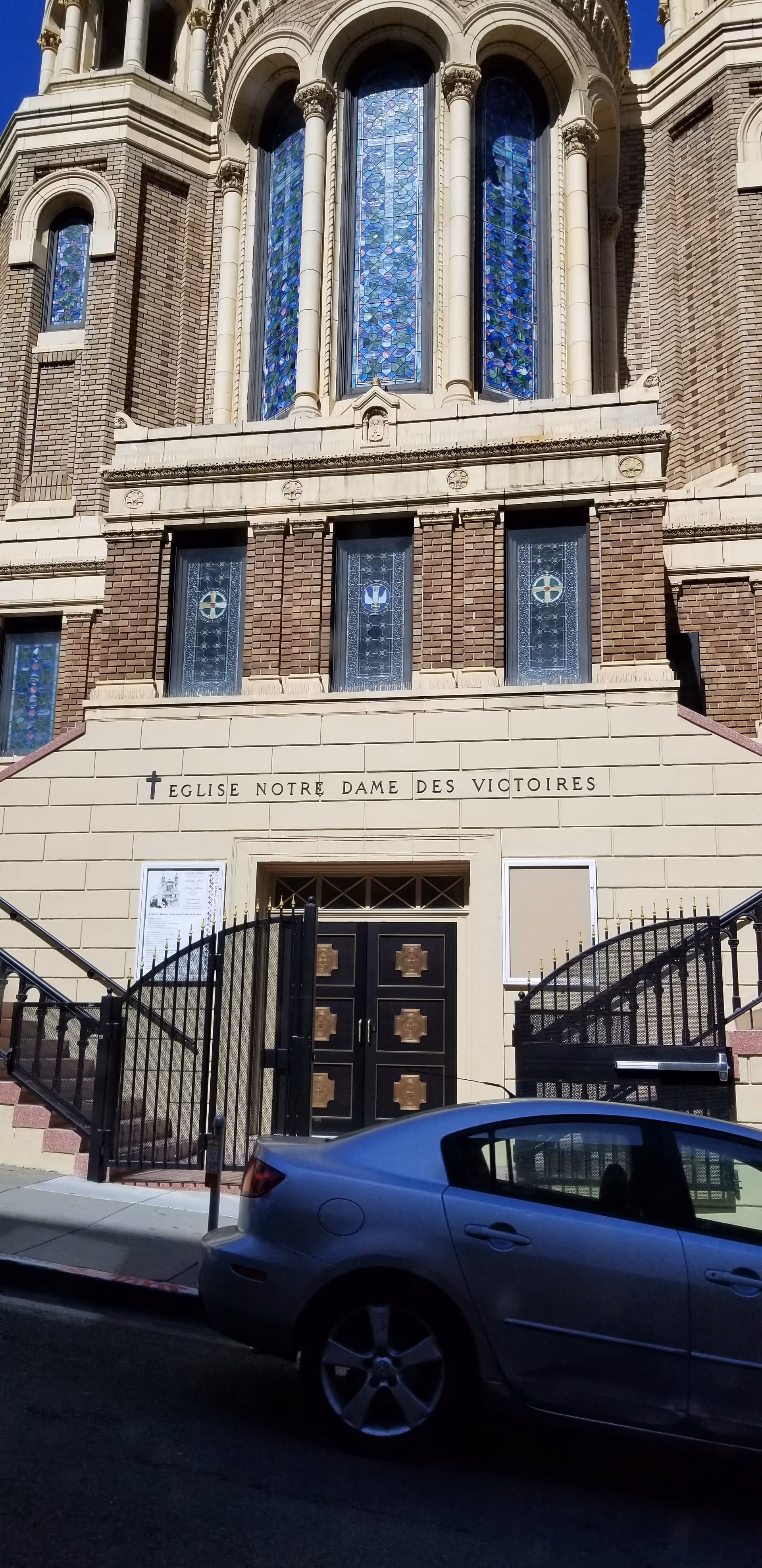 Ornate church facade with arched stained-glass windows, stone detailing, and a blue car parked outside.