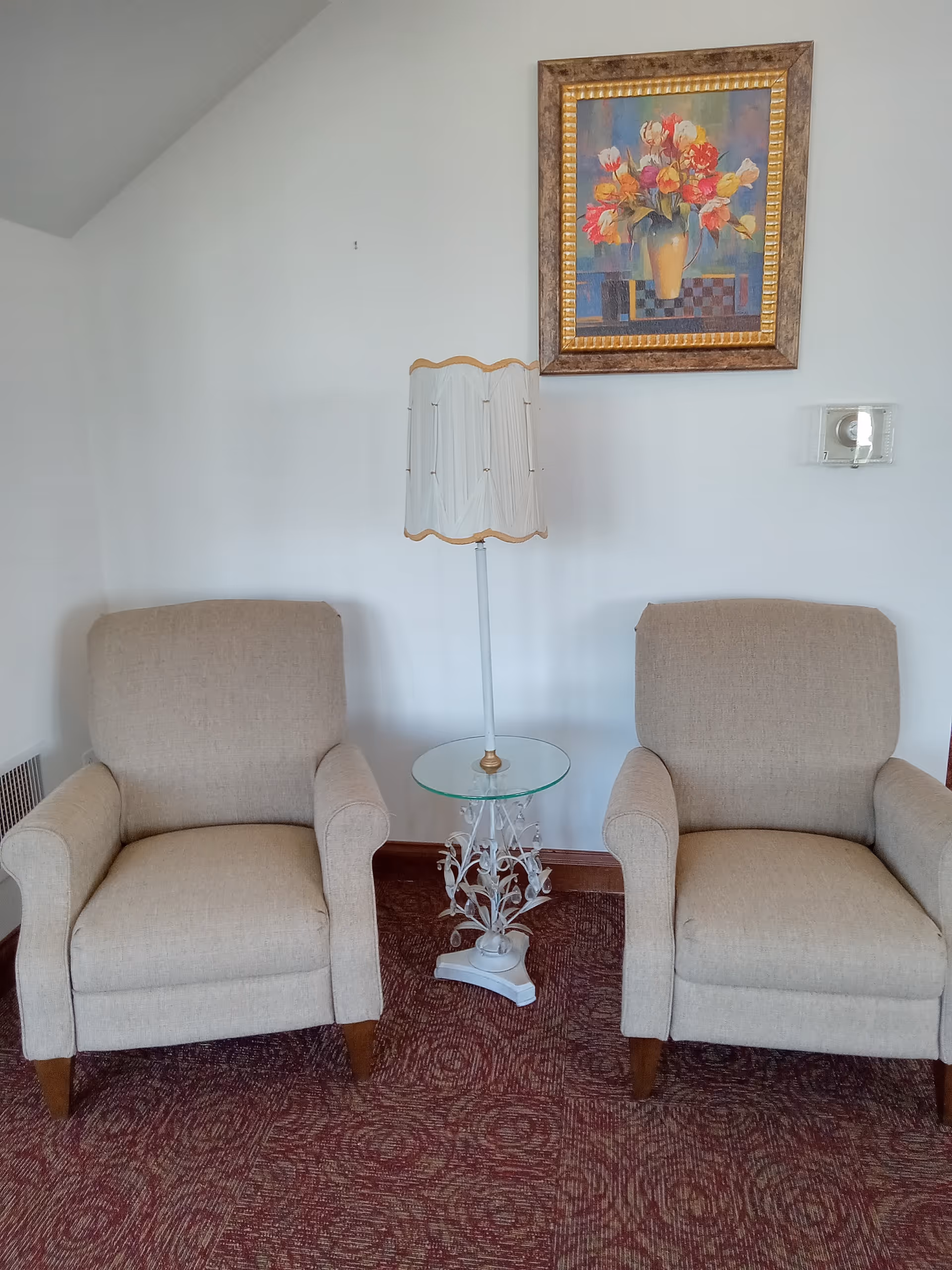 Two beige armchairs flank a glass-topped table with ornate base, lamp, and framed floral painting.