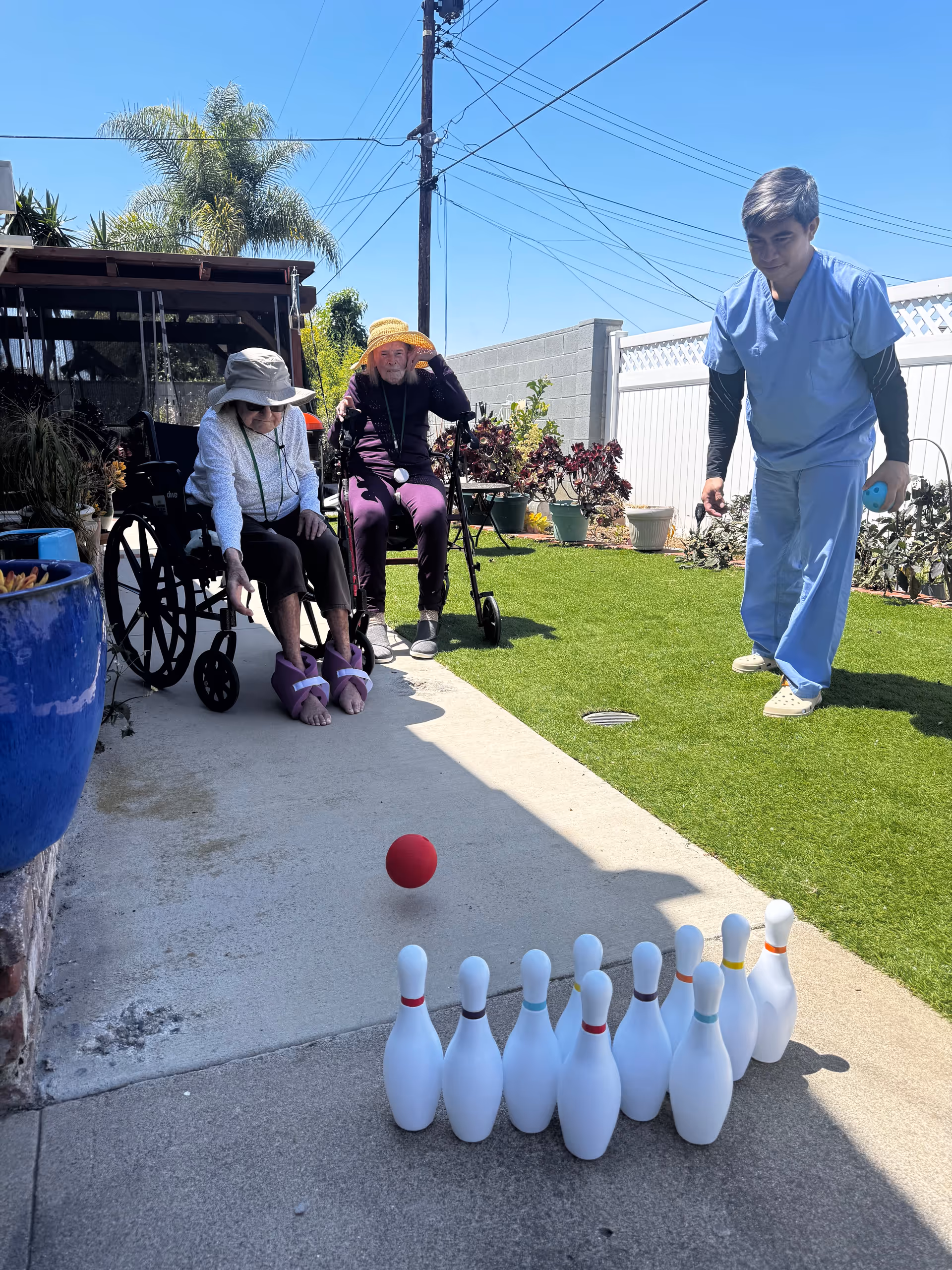 Elderly residents in wheelchairs join a caregiver for an outdoor lawn bowling game.