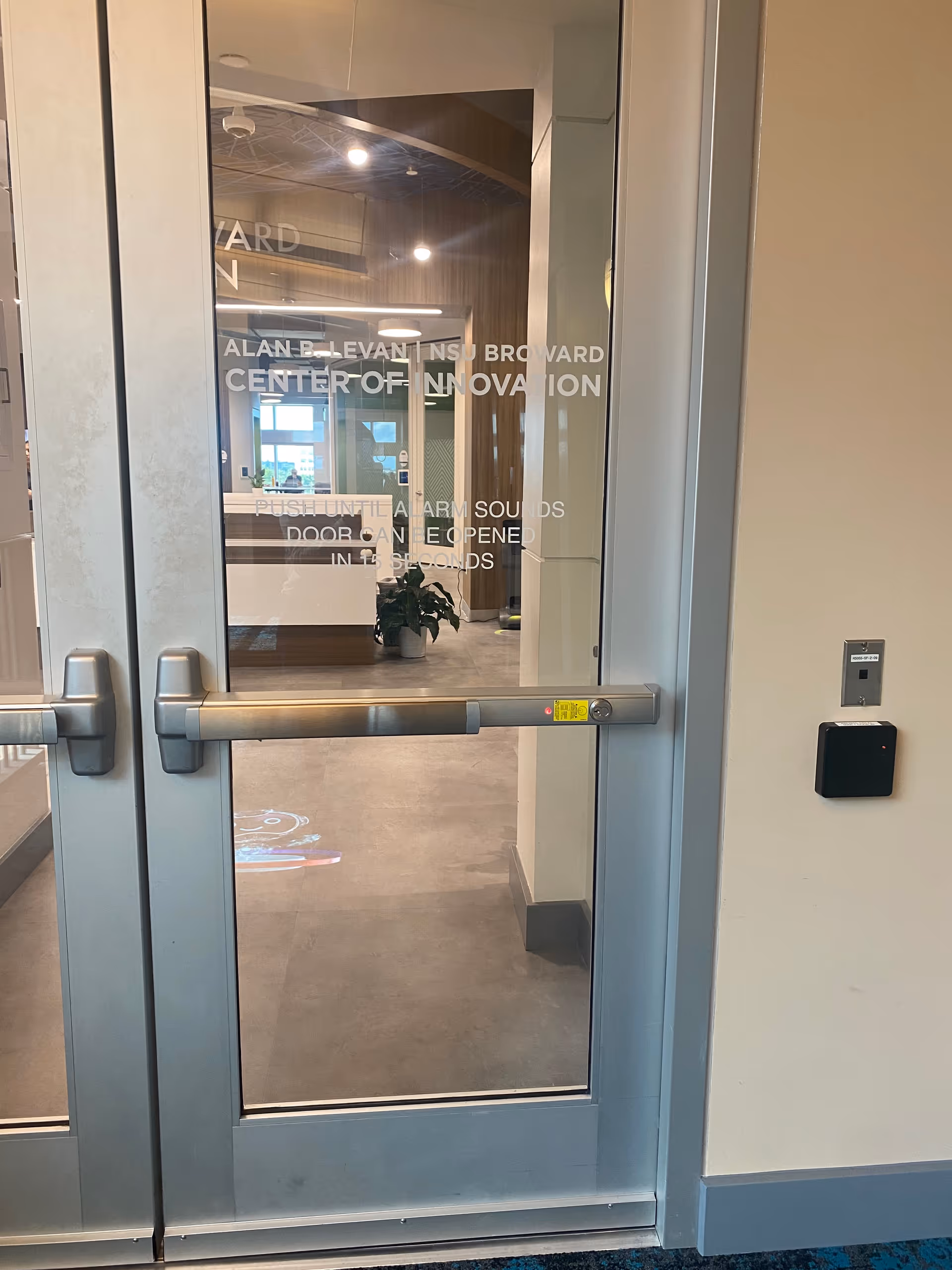 Glass doors open to a modern interior hallway with a plant and signage.