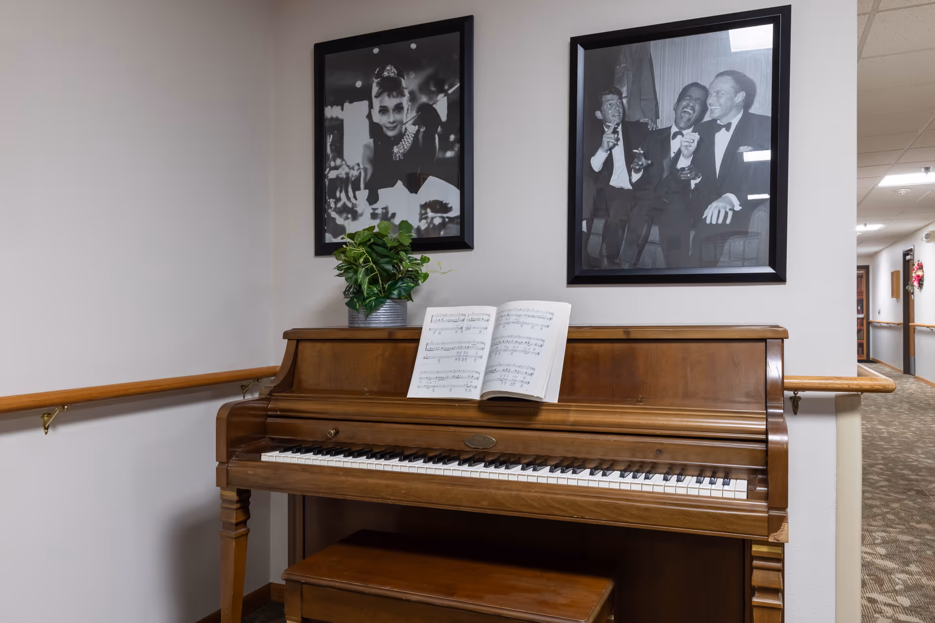 A wooden upright piano with an open sheet of music on the stand and a small potted plant on top. Above the piano are two black and white framed photographs, one of a woman and the other of three men laughing. The piano is placed against a wall in a hallway with handrails and carpeted flooring.
