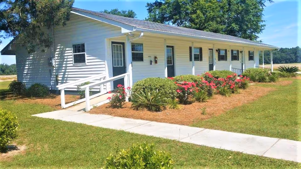 Single-story white building with multiple doors and windows, surrounded by a well-maintained garden with green bushes and pink flowers, under a clear blue sky.