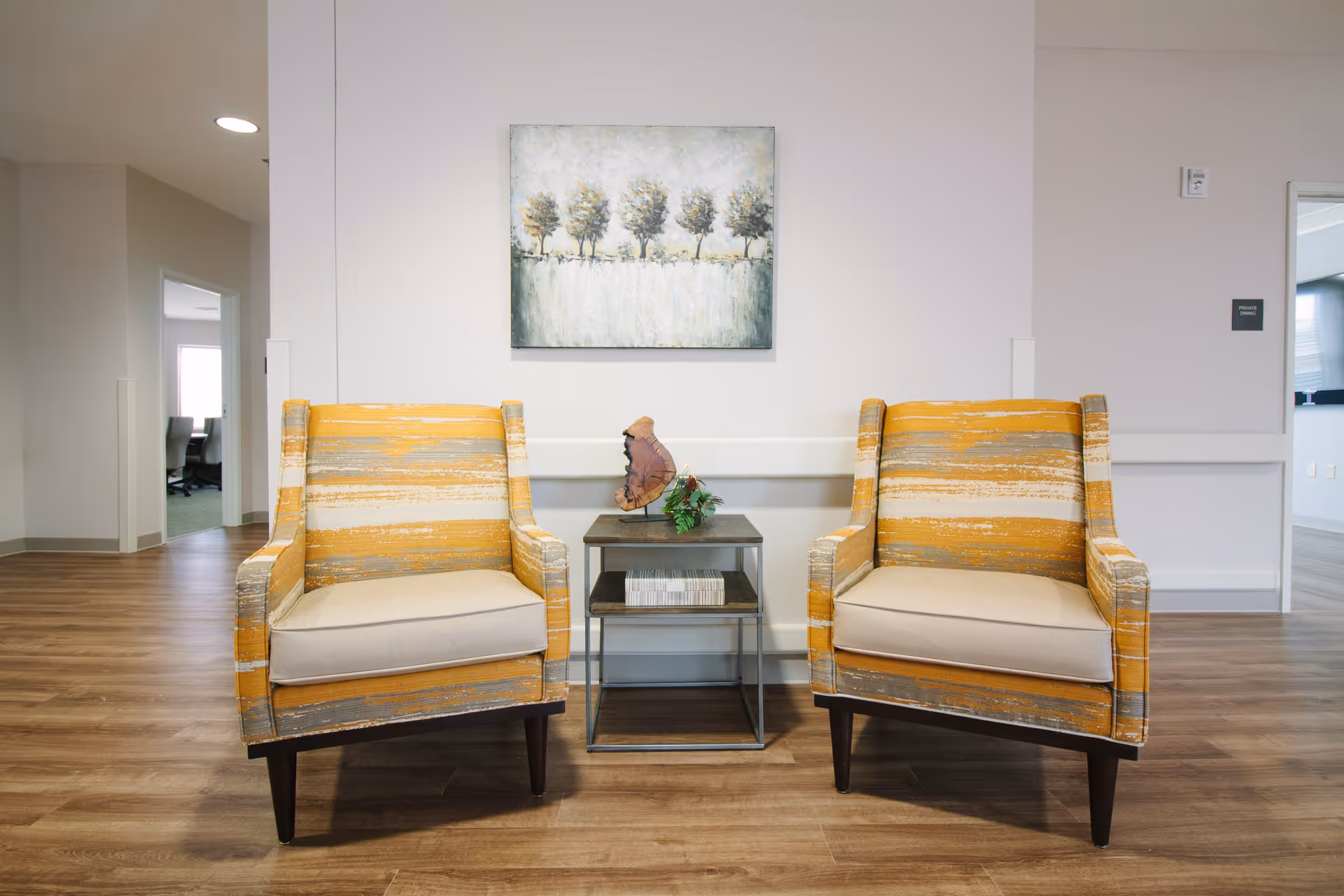 Two striped orange chairs flank a small table beneath a landscape painting in a modern lobby.