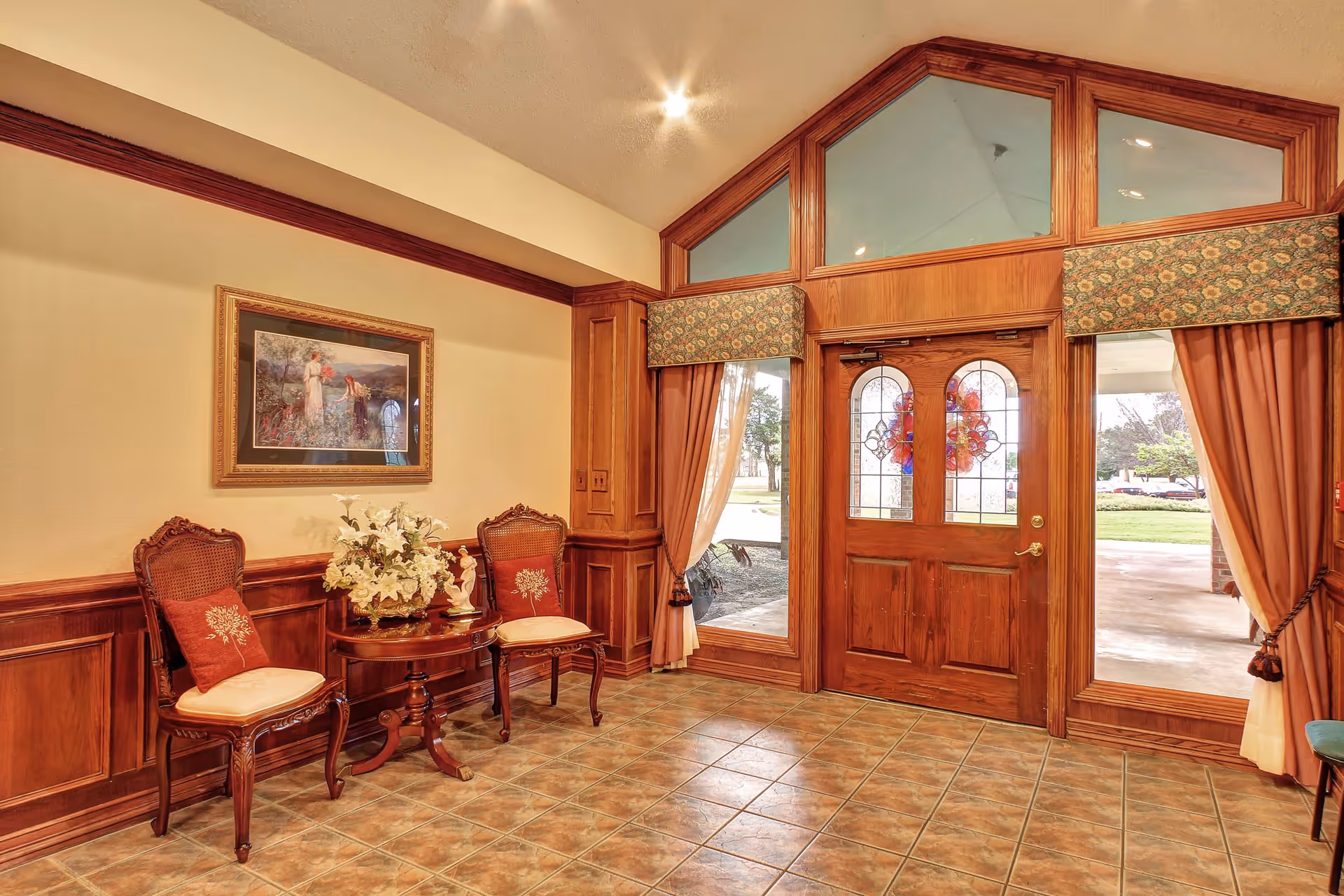 Entrance area of Crestwood Health and Rehabilitation Center featuring a wooden double door with decorative glass panels, flanked by large windows with floral valances and curtains. Two ornate wooden chairs with cushions and a small round table with a floral arrangement are positioned against a wall with wood paneling and a framed painting above.