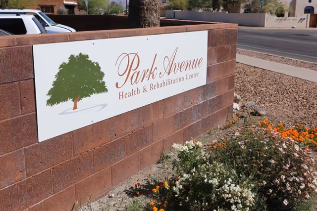 A white sign mounted on a low brick wall that reads 'Park Avenue Health & Rehabilitation Center' with flowers and a street visible behind it.