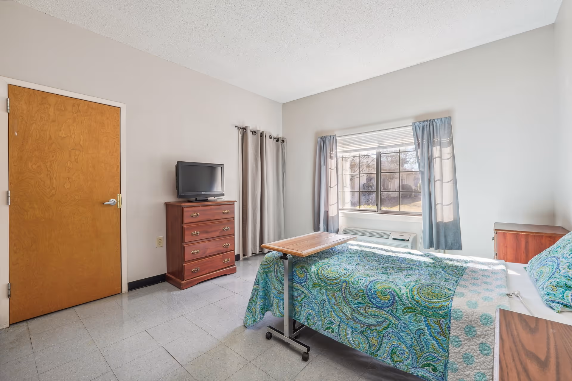 Senior living bedroom with a bed, dresser, small table, window, and TV.