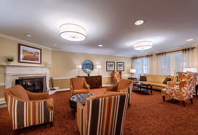 A cozy senior living facility common area with a fireplace on the left wall, surrounded by four armchairs with striped and solid upholstery arranged around a round tufted ottoman. There is a beige sofa and two floral-patterned armchairs near large windows with sheer curtains. The room has warm lighting from ceiling fixtures and table lamps, with framed artwork and a round mirror on the walls.