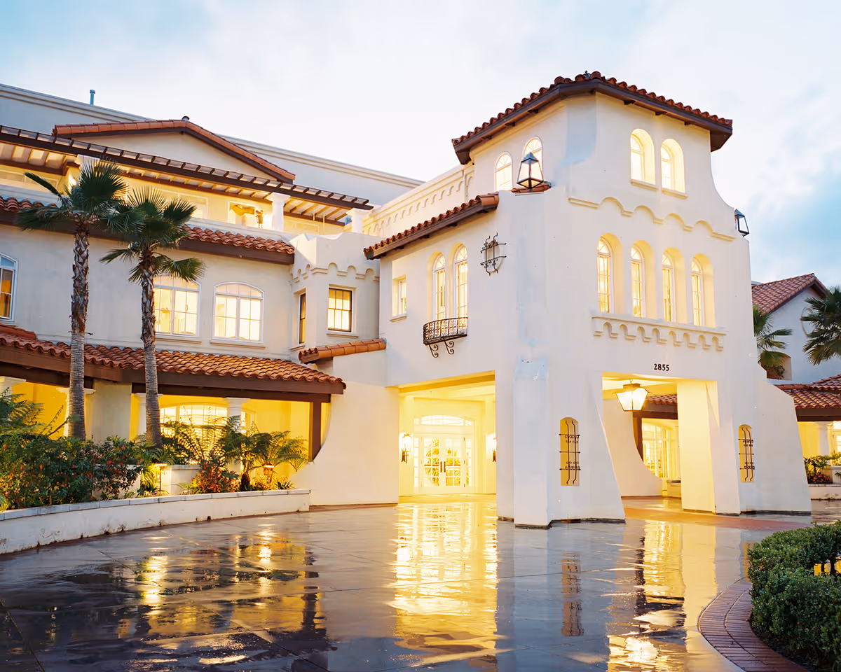 Exterior view of a white Mediterranean-style senior living facility with palm trees and a covered entry.