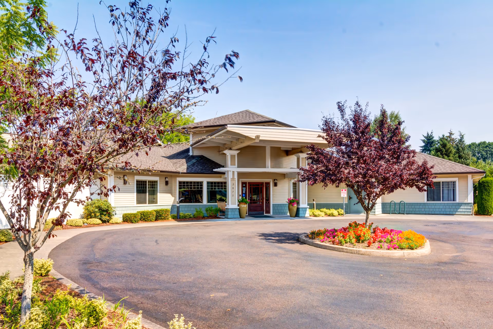 Exterior view of Timberwood Court Memory Care facility showing a single-story building with a covered entrance, surrounded by landscaped trees and a circular flower bed in front of the driveway.