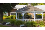 A white gazebo sits in a landscaped outdoor area with a building behind.