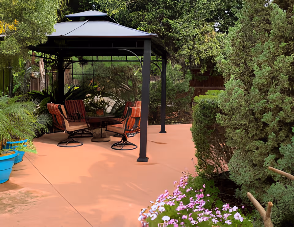 Outdoor patio area with a black metal gazebo covering four cushioned chairs arranged around a small round table. The patio is surrounded by lush green trees, bushes, and flowering plants in a garden setting.