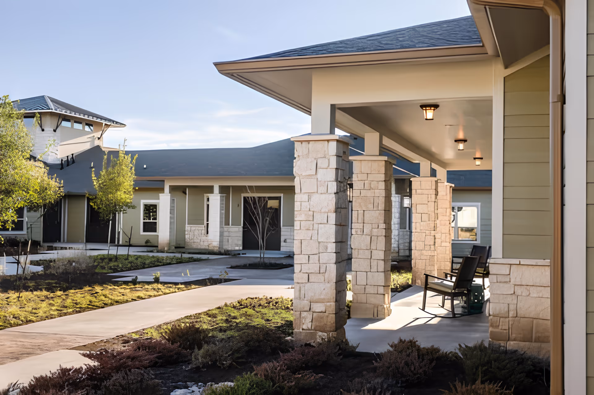 Covered entry and courtyard of a senior living facility with stone columns, a walkway, and rocking chairs on a porch.