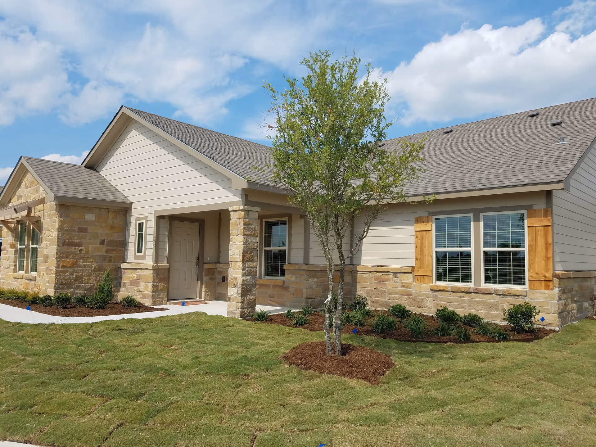 One-story assisted living facility exterior with stone accents, beige siding, entryway, and landscaped lawn.