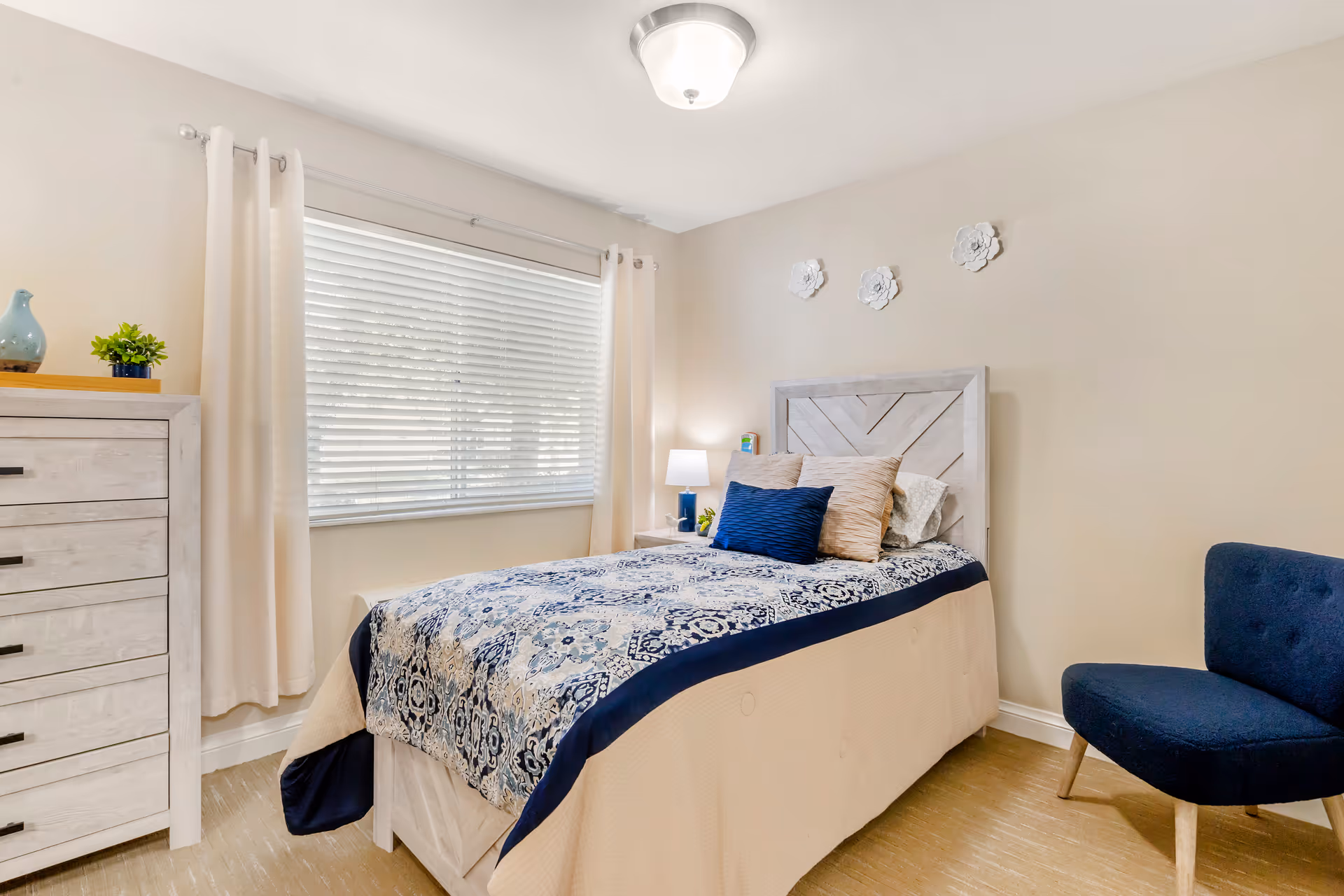 Cozy bedroom with a bed, dresser, window with blinds, and a blue chair.