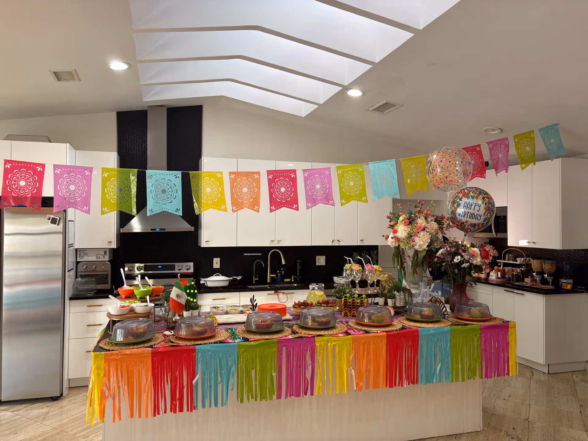Colorful birthday party setup in a bright kitchen with a long decorated table and balloons.