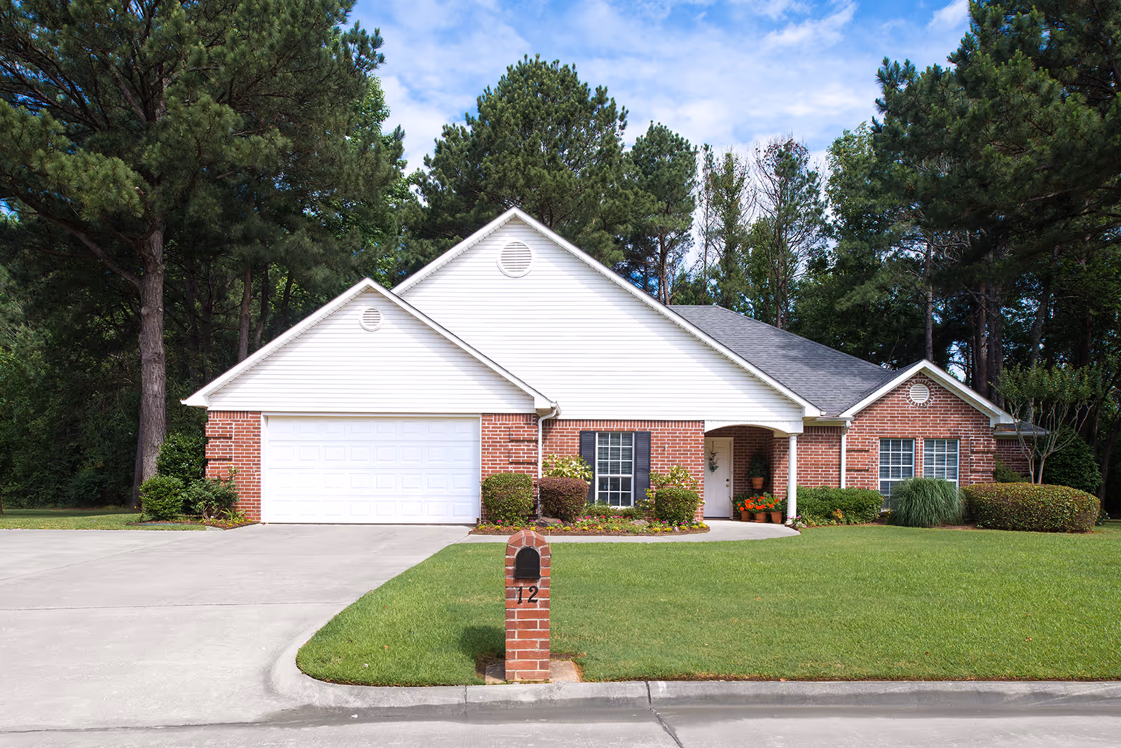Front exterior view of a single-story brick and white siding house with a two-car garage, a well-maintained lawn, shrubs, and trees in the background under a partly cloudy sky.