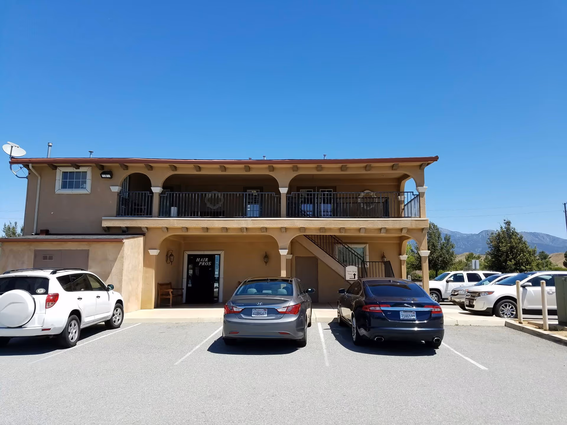 Two-story beige building with a balcony, stairs, and several parked cars in front.
