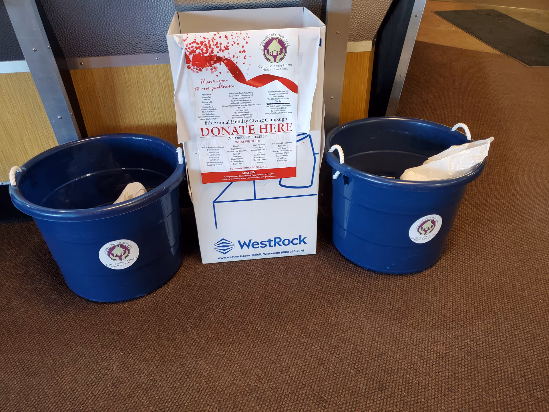 A donation box sits between two blue recycling bins on a brown carpet.