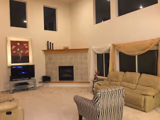 Spacious living room with beige carpet, a beige sectional sofa, a patterned armchair, a fireplace with a wooden mantle, a TV on a stand, and large windows near the ceiling.